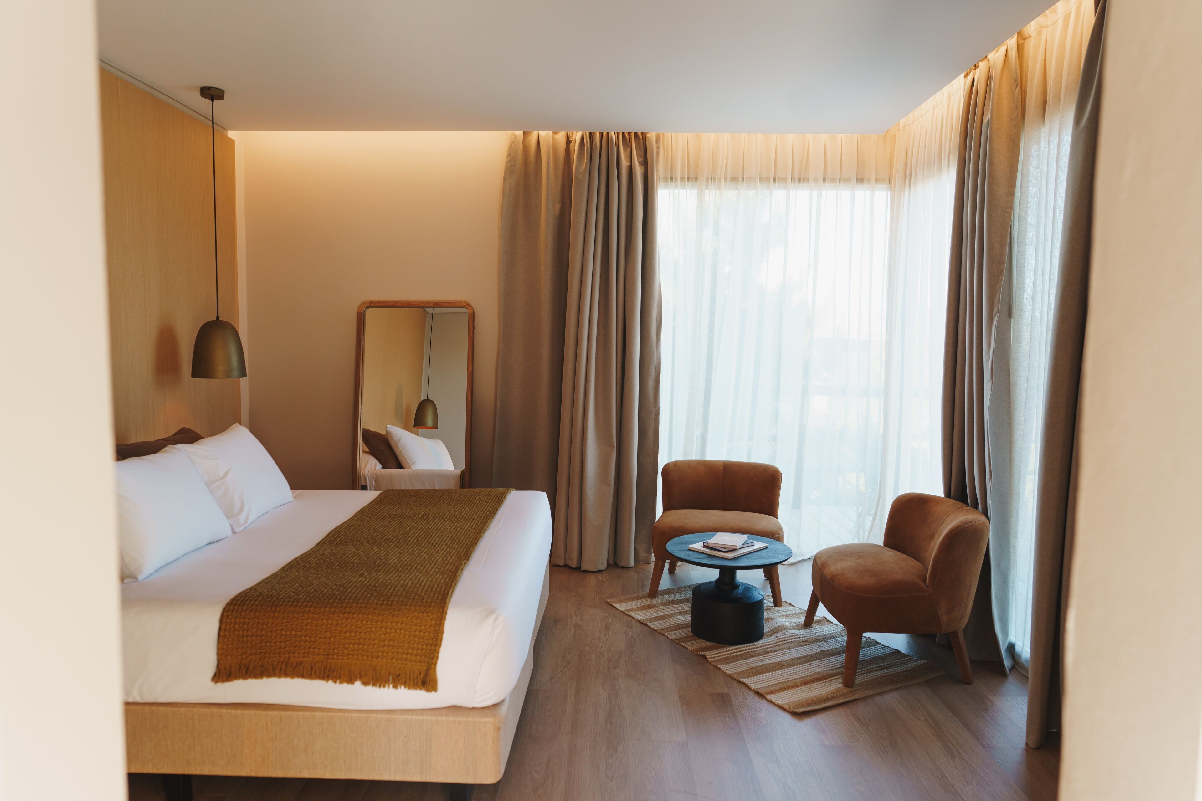 A hotel room with a large bed, a round coffee table, a chair, a mirror, and a window with curtains.