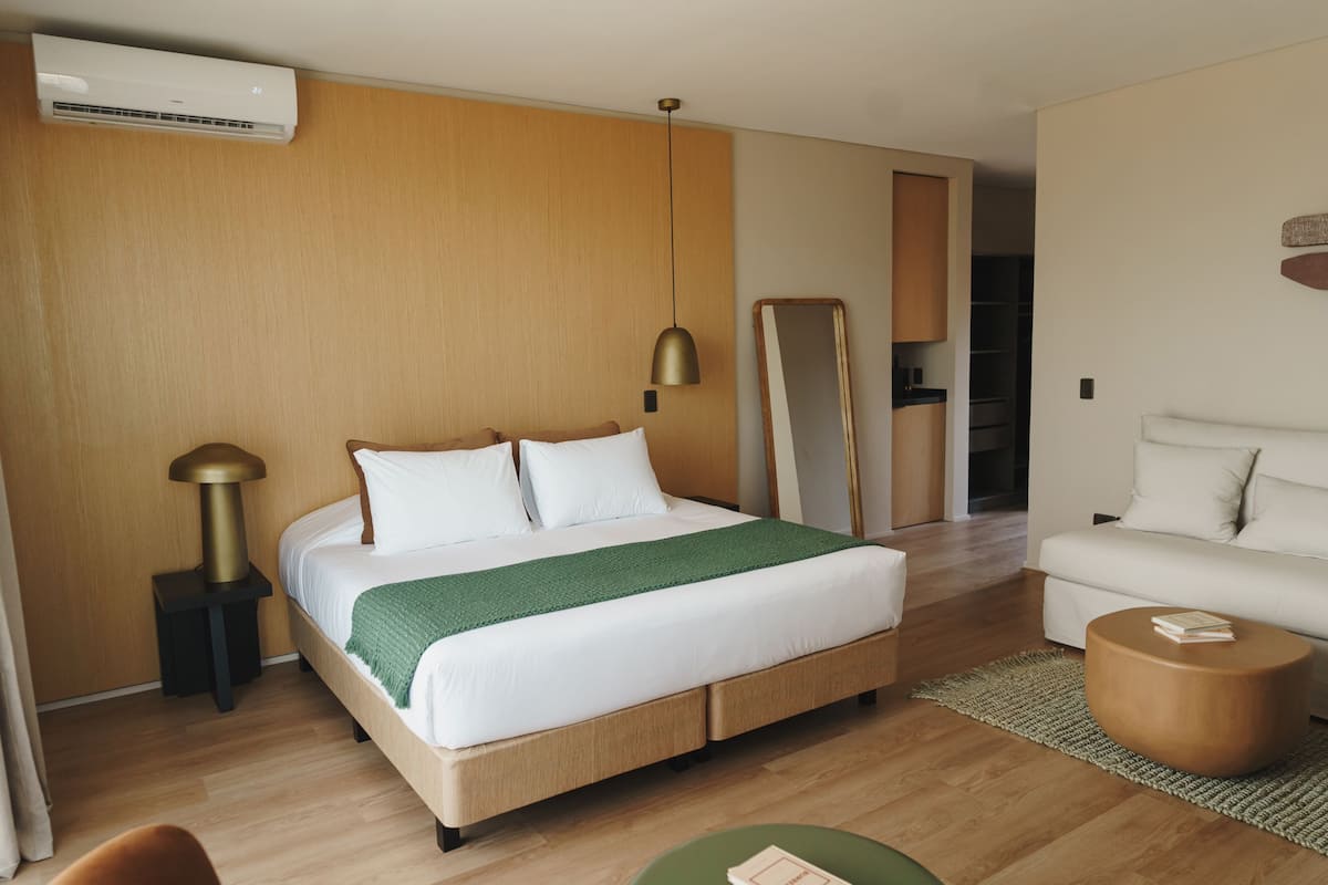 A modern hotel room with a wooden floor, a bed with white bedding, a green throw, a round wooden side table, a white sofa, and a large mirror.