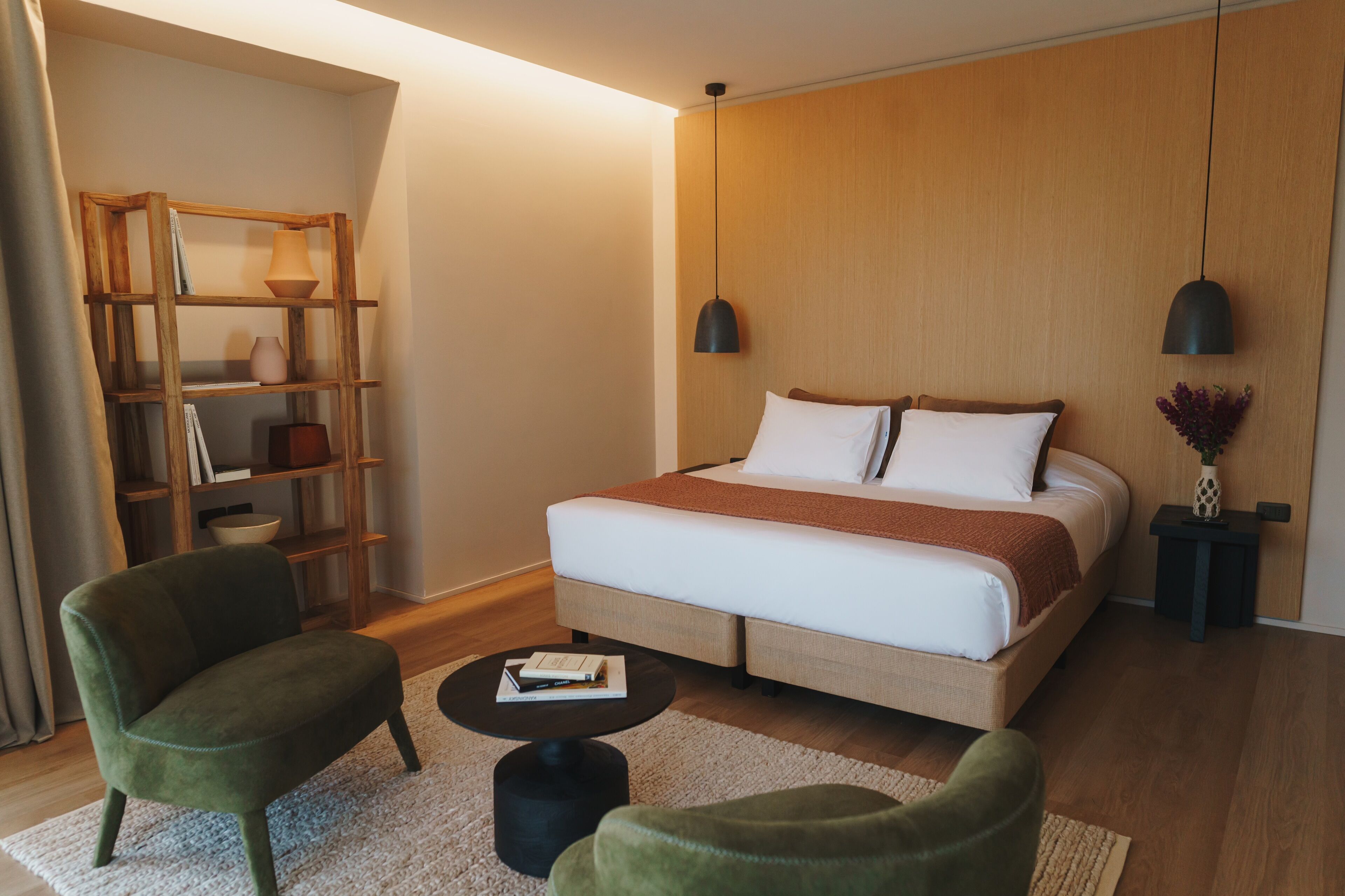 Una habitación de hotel con una cama grande, dos mesitas de noche, una estantería, un sillón verde y una pequeña mesa redonda.