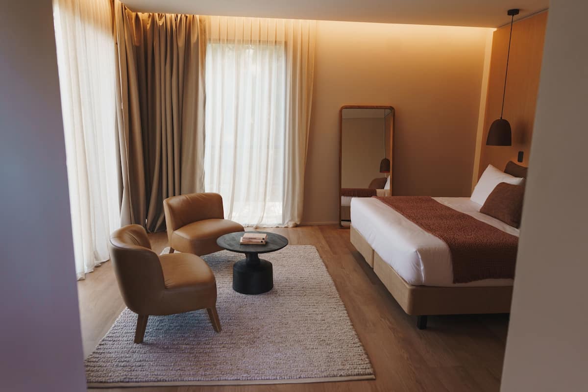 A hotel room with a bed, two armchairs, a small round table, a large mirror, and a hanging light fixture.