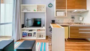 A compact living space with a kitchenette, a flat-screen TV, a small bar area, and a window with curtains. - Maximus Luxury in Nyc Berrini (São Paulo)