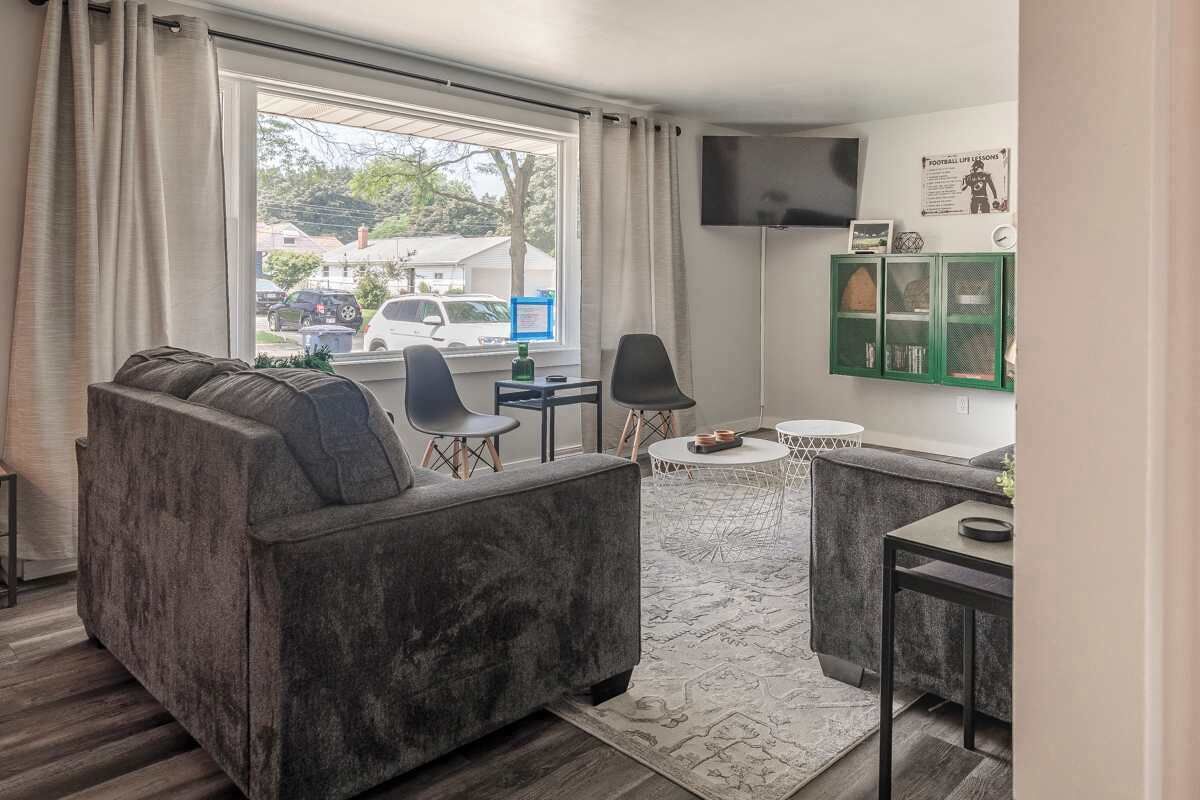 Ein modernes Wohnzimmer mit einem grauen Sofa, zwei Sesseln, einem kleinen runden Tisch und einem an der Wand befestigten Fernseher.
