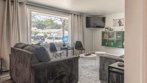 Ein modernes Wohnzimmer mit einem grauen Sofa, zwei Sesseln, einem kleinen runden Tisch und einem an der Wand befestigten Fernseher.