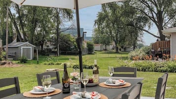 Ein Essbereich im Freien mit einem Tisch für zwei Personen unter einem großen Sonnenschirm, umgeben von einem grünen Rasen und Bäumen.
