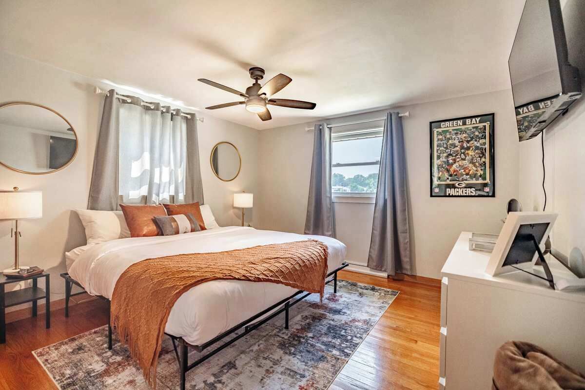 Ein Schlafzimmer mit Bett, Deckenventilator, Schreibtisch, Spiegel und Fenster mit Vorhängen.