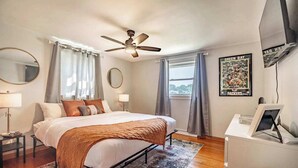 Ein Schlafzimmer mit Bett, Deckenventilator, Schreibtisch, Spiegel und Fenster mit Vorhängen.