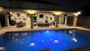 Una piscina con una terrazza, un'area salotto coperta e una casa sullo sfondo.