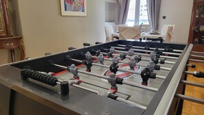 A foosball table in a room with a painting on the wall, a window with curtains, and a table with chairs in the background. - Poseidon Executive Suite in the heart of Athens Riviera. Edem beach and marina. (Paleo Faliro)