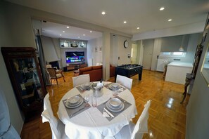 A dining area with a round table set for six, a television in the background, and a kitchen area visible through an open door.