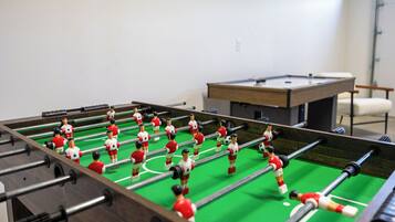Une table de baby-foot avec des figurines rouges et blanches, dans une pièce où se trouvent également une table de billard et une chaise.