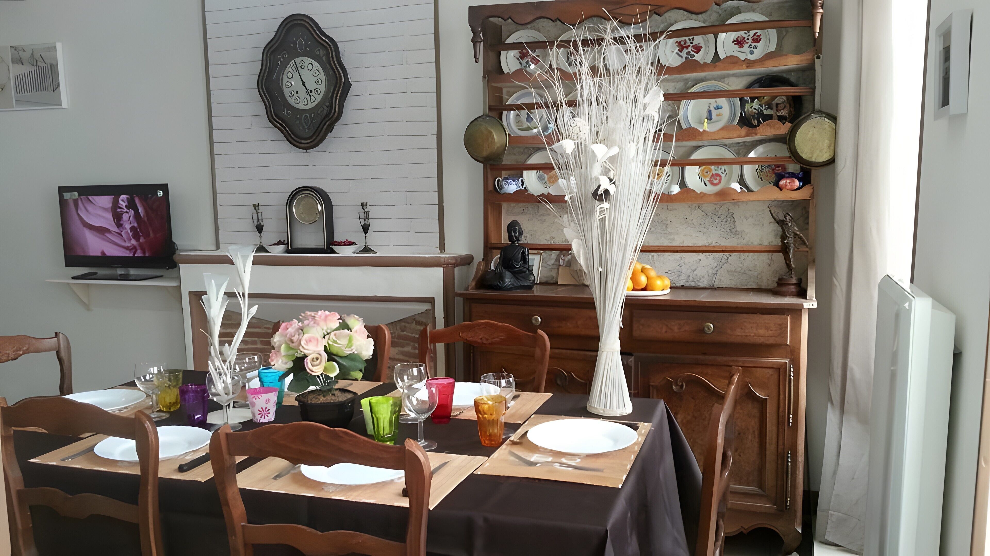 A dining room with a table set for four, a clock on the wall, and a shelf with dishes and a Buddha statue.