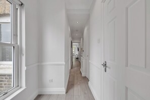 A hallway with white walls, wooden flooring, and a window on the left. - Beaufort Gardens Collection (London)