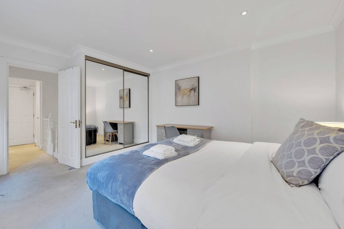 A bedroom with a bed, a desk, a chair, a mirror, and a door leading to another room.