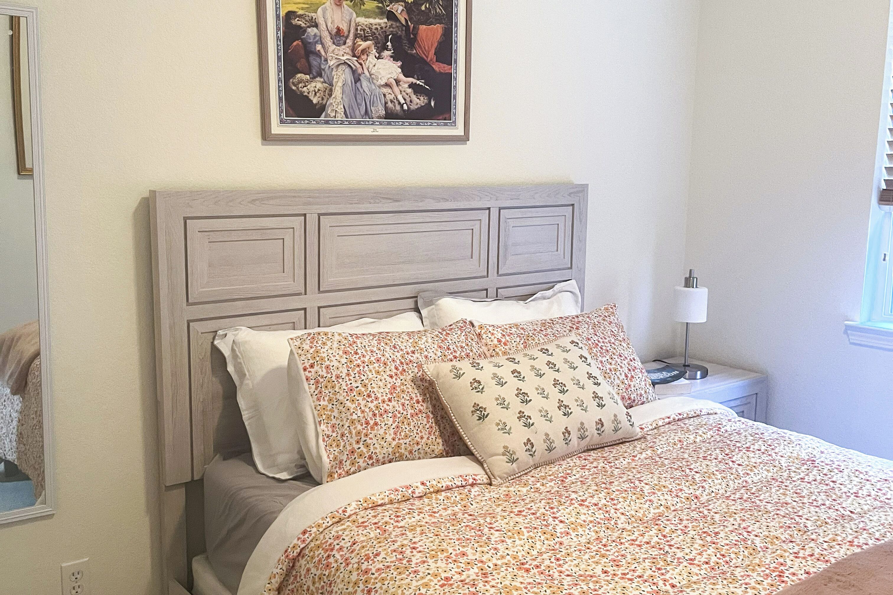 Een slaapkamer met een houten hoofdbord, beddengoed met bloemenprint, een nachtkastje met een lamp, en een ingelijst schilderij aan de muur.