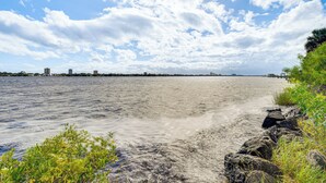 Ein Strand mit felsiger Küste, ruhigem Wasser und einer in der Ferne liegenden Stadtansicht unter bewölktem Himmel.