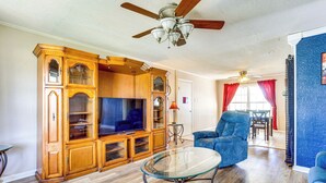 Un salon avec un meuble de rangement en bois pour les appareils multimédias, une table basse en verre, un fauteuil bleu et un ventilateur de plafond.