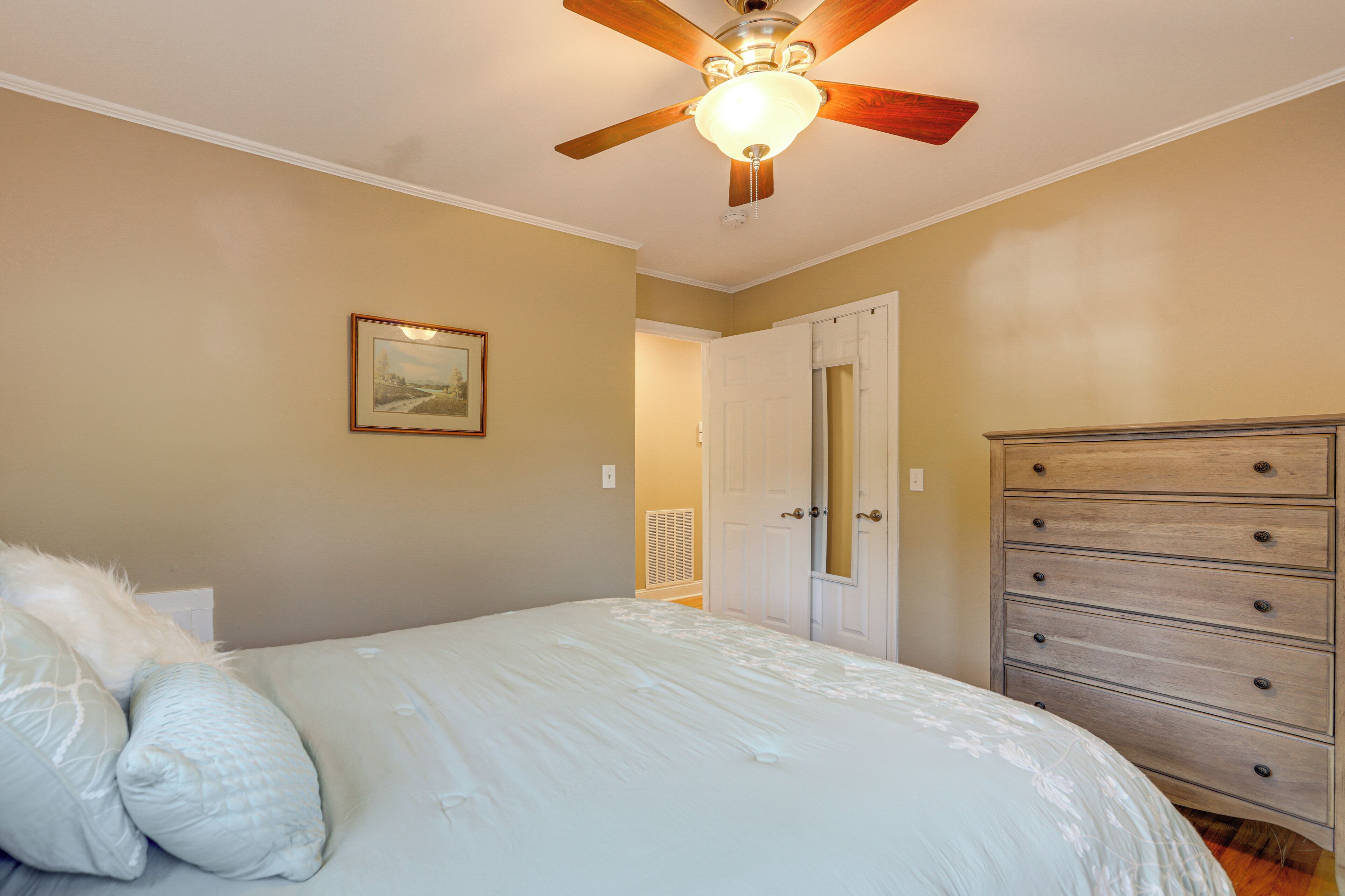 A bedroom with a bed, a dresser, a ceiling fan, and a door leading to another room.