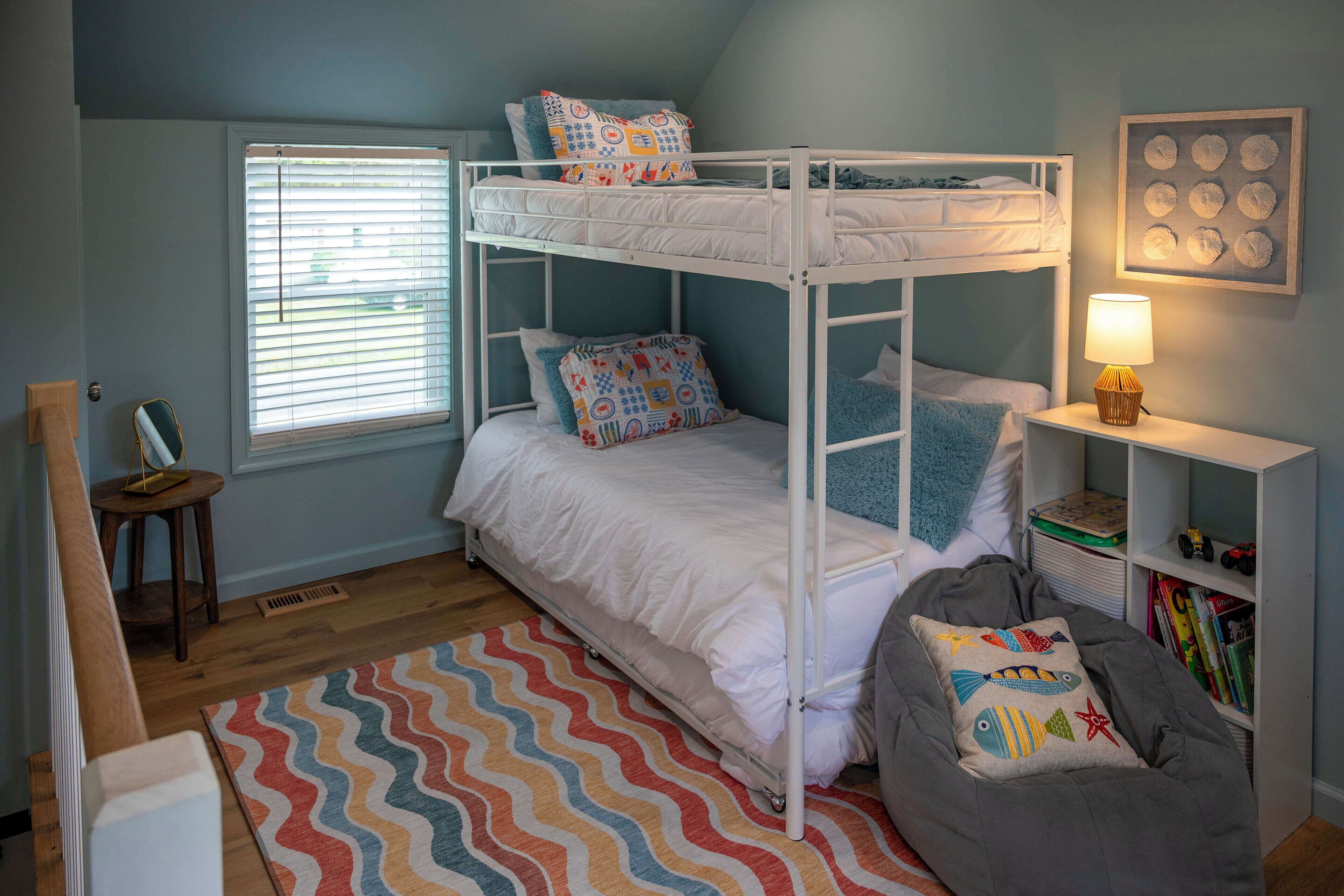 A bunk bed room with a window, a bedside lamp, a bookshelf, and a colorful rug.