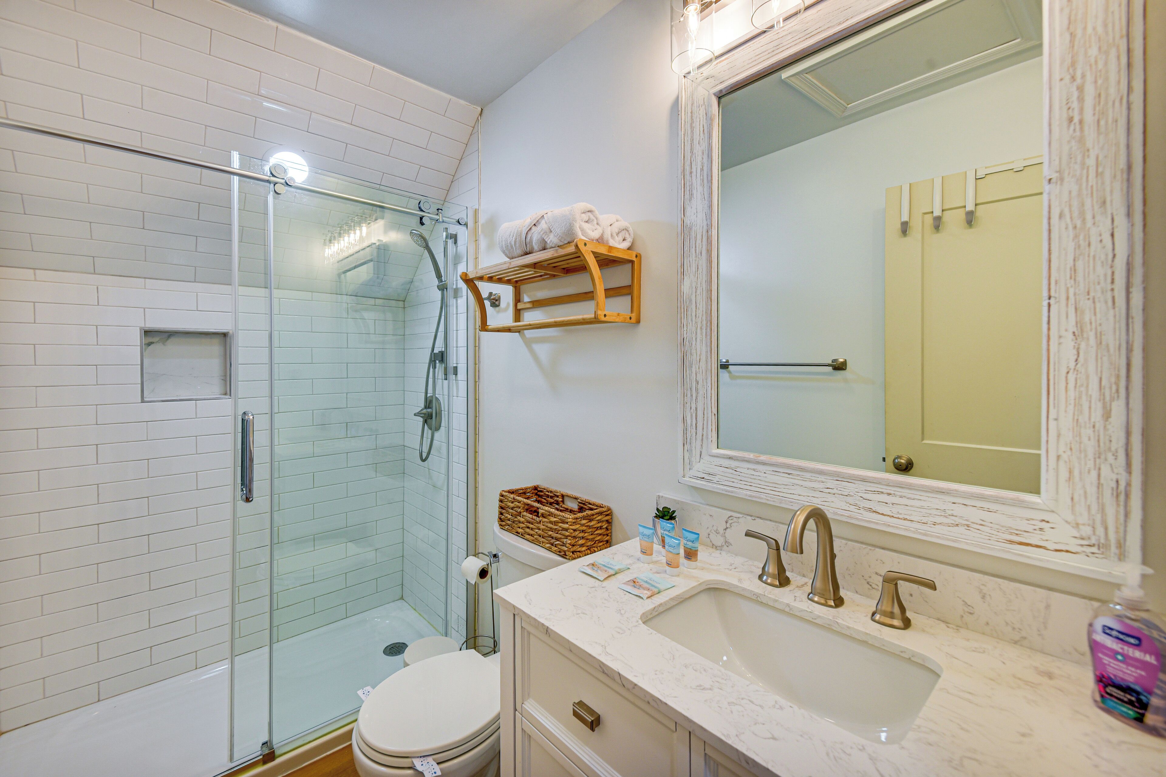 A bathroom with a glass shower enclosure, a white toilet, a marble sink, and a wooden shelf with towels.