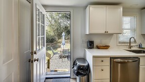 A kitchen with white cabinets, a stainless steel dishwasher, and a view through the open door to an outdoor seating area. - Beach Days & Dtwn Charm! Cape Charles Cottage (Cape Charles)
