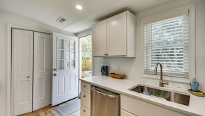 A modern kitchen with white cabinets, a stainless steel dishwasher, a sink with a faucet, and a window with blinds. - Beach Days & Dtwn Charm! Cape Charles Cottage (Cape Charles)