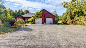A red barn with two white garage doors, surrounded by trees and a gravel driveway. - Stars & Sunsets! Door Peninsula Retreat w/ Deck (Egg Harbor)