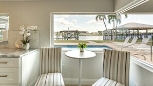 A room with a view of a pool and outdoor seating, a small table with a plant, and striped chairs.