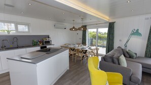 A modern kitchen with white cabinets, a central island, and a dining area with wooden chairs. - Dahlia (Redruth)