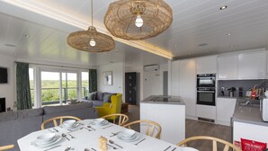 A modern kitchen with white cabinets, a built-in oven, and a dining area with a white table set for four. - Dahlia (Redruth)