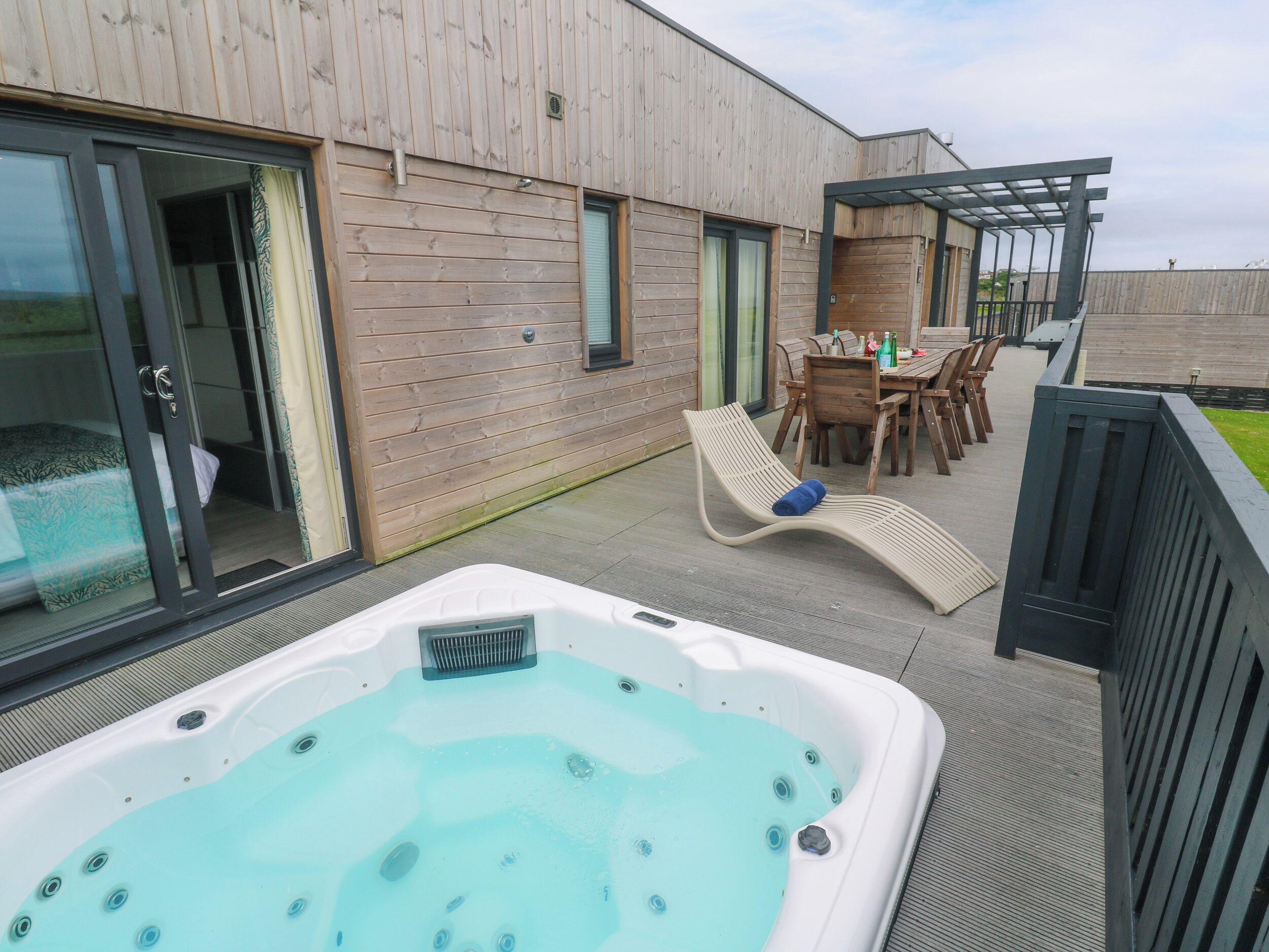 Ein Whirlpool auf einer Terrasse mit Holzdeck, ein Essbereich mit Stühlen und Tisch sowie eine Glastür zu einem Raum.