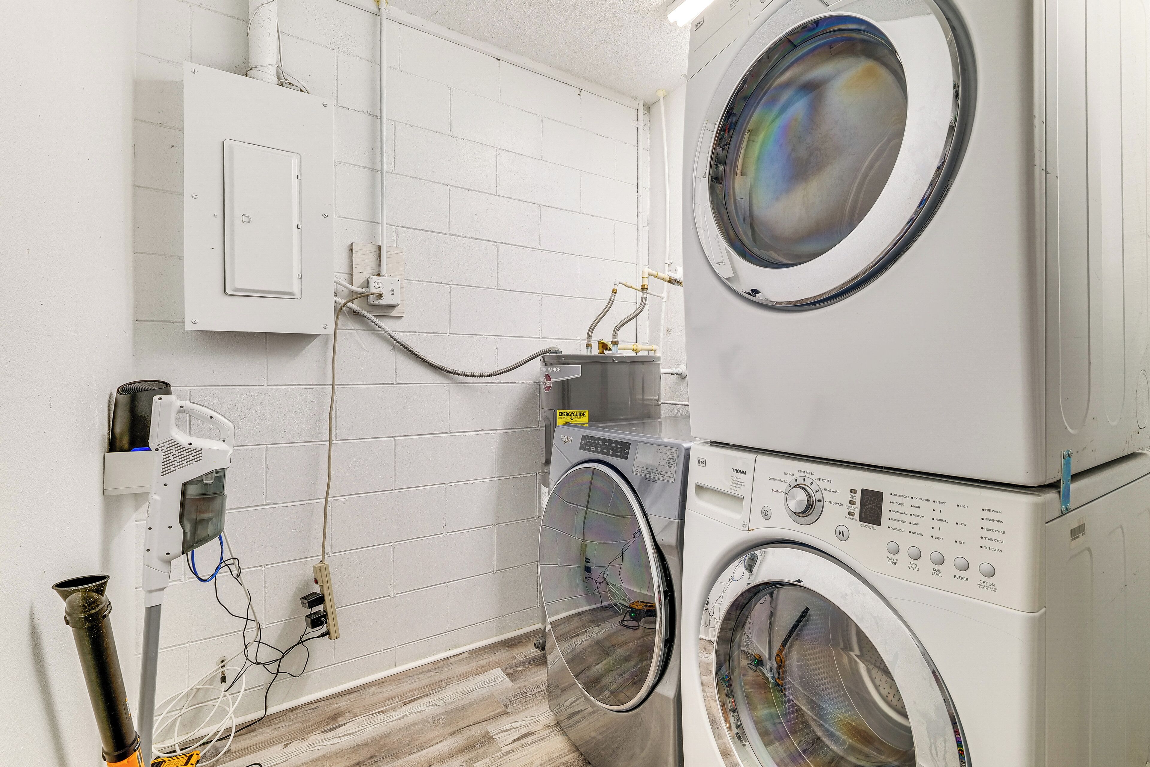 A laundry room with two stacked front-loading washing machines, a utility room with a white brick wall, and visible electrical outlets and wiring.