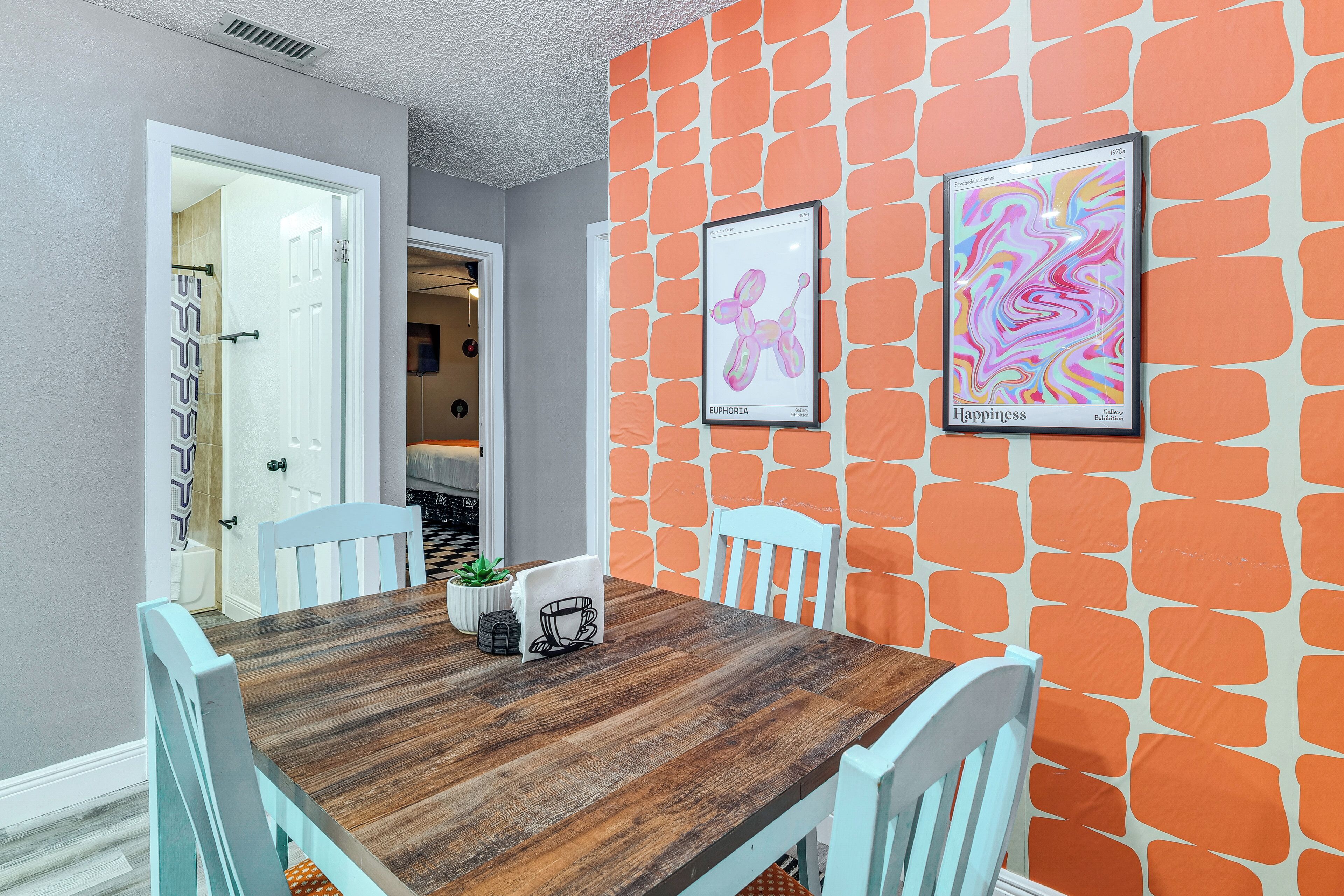 Un espace repas avec une table en bois, des chaises blanches et un mur orné de motifs géométriques orange.