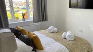 A hotel room with a bed, a bedside table with a lamp, a TV mounted on the wall, and a window with curtains. - Stunning Renovated Three Bedroom House (Poole)