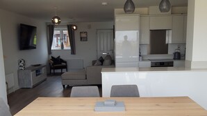 A modern living room with a wooden dining table and grey upholstered chairs, a grey sofa, and a flat-screen TV mounted on the wall. - Stunning Renovated Three Bedroom House (Poole)