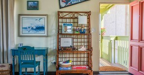 A room with a wooden chair, a small table, a wicker shelf with books and decorative items, and a framed picture on the wall.