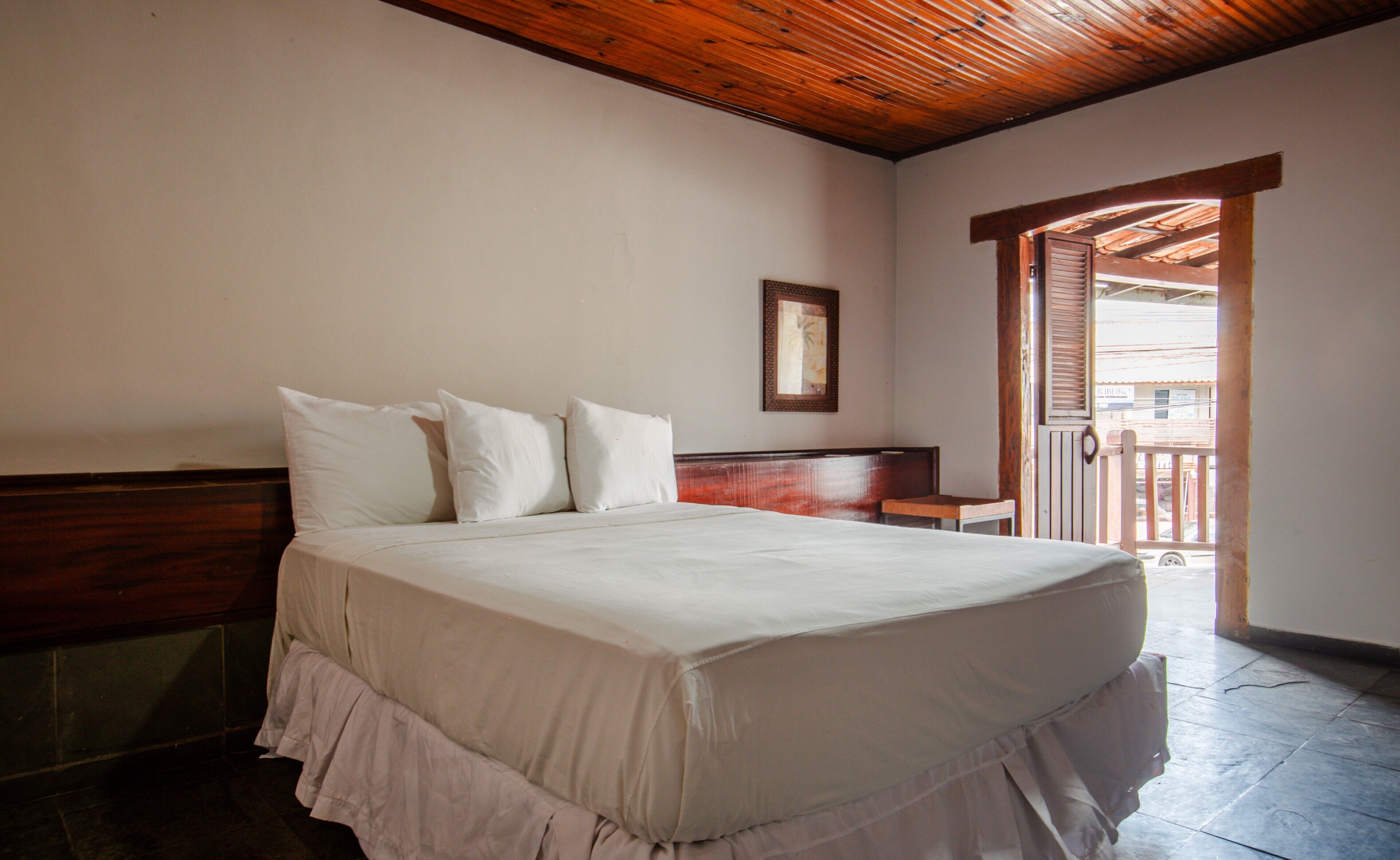 Quarto com cabeceira de madeira, cama com roupa de cama branca, uma mesinha e porta para varanda.