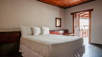 Quarto com cabeceira de madeira, cama com roupa de cama branca, uma mesinha e porta para varanda.