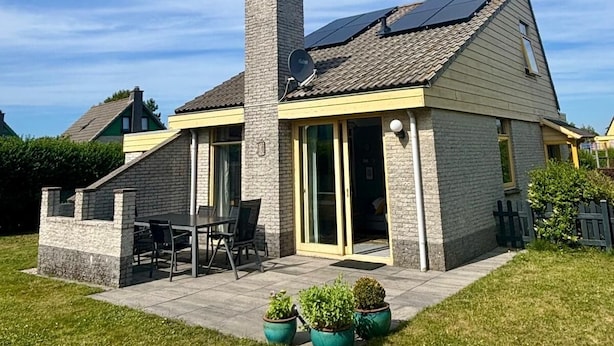 Une maison avec une cheminée en pierre, des panneaux solaires, une terrasse avec des chaises et des plantes en pot.