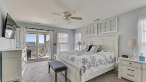 A bedroom with a large bed, a ceiling fan, a desk with a lamp, and a view of the outside.
