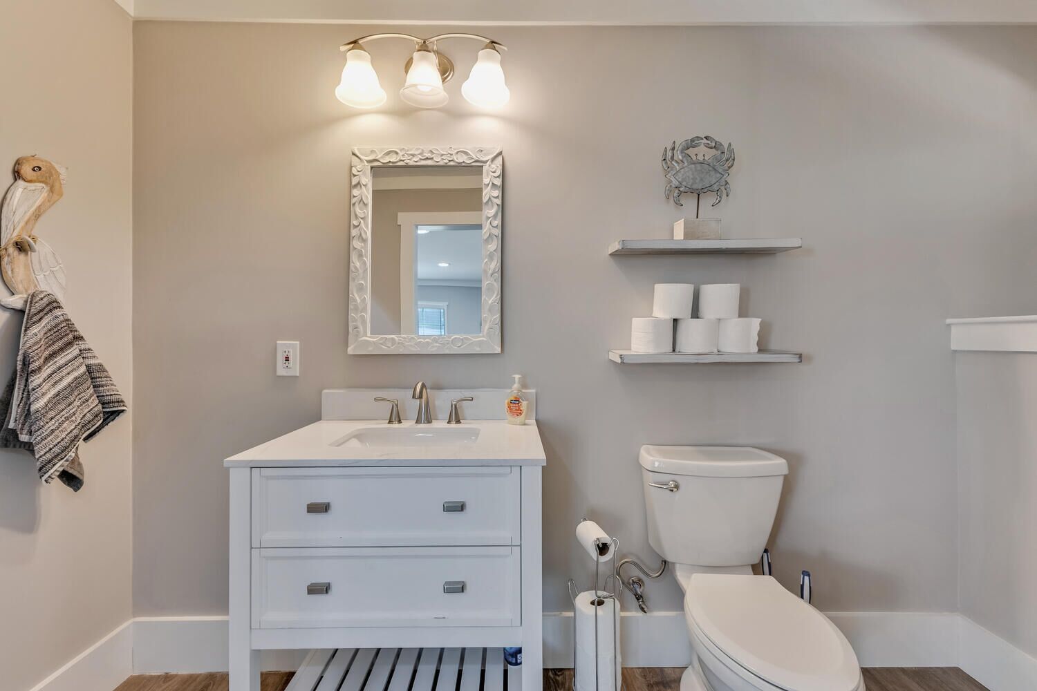 A bathroom with a white vanity, a mirror, a toilet, and a shelf with toilet paper.