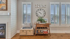 A room with a fireplace, a clock, a 'Relax' sign, and a wooden shelf with books and a plant.