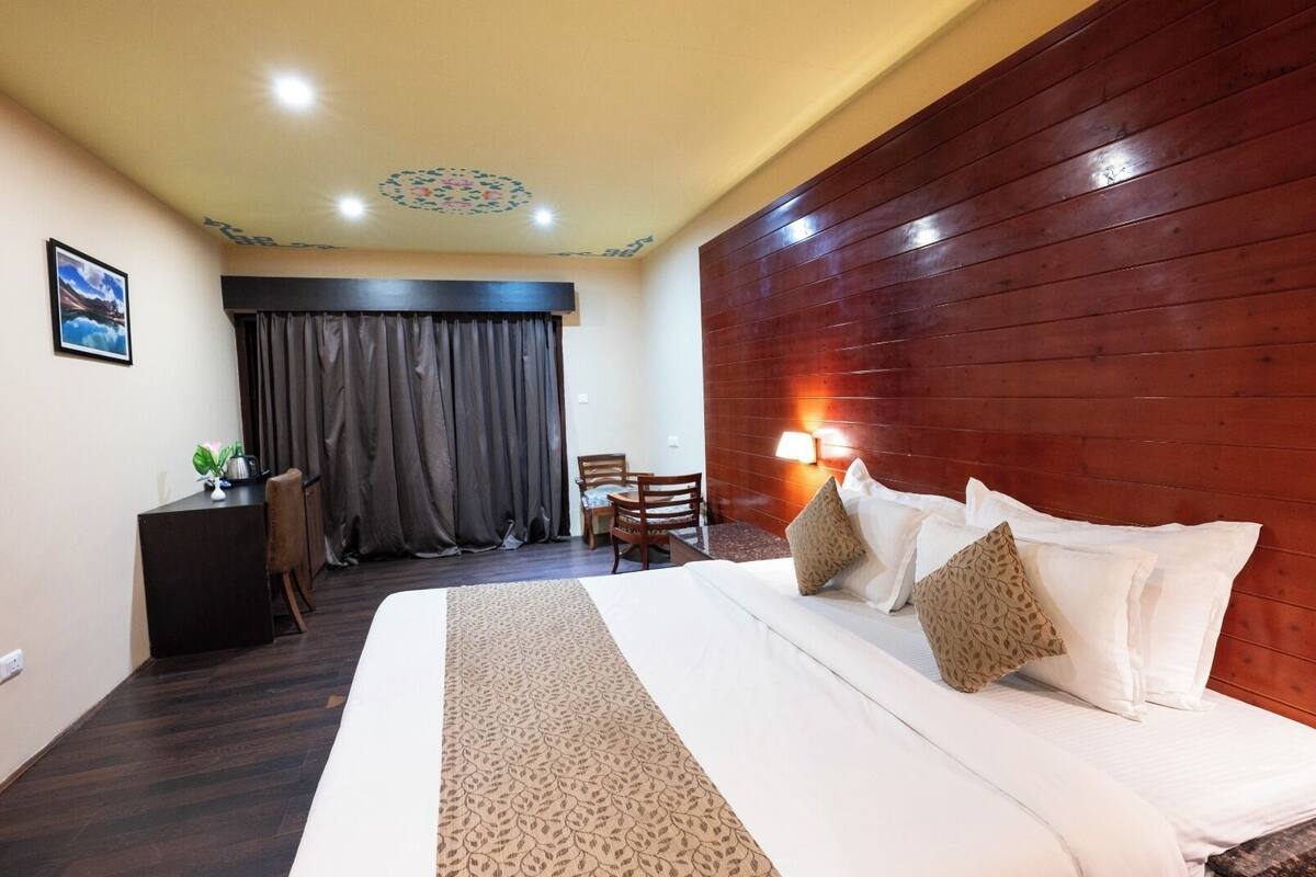 Een hotelkamer met een groot bed, een houten hoofdeinde, een bureau met stoel, een klein tafeltje met stoelen en een raam met gordijnen.