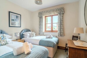 A bedroom with two beds, a wooden nightstand, a desk with a lamp, and a window with curtains.