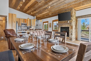 A dining area with a long wooden table set for a meal, a stone fireplace, and a kitchen with wooden cabinets.