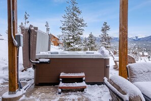 A hot tub with a wooden deck, surrounded by snow-covered trees and a mountain view.
