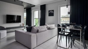 A modern living room with a grey sofa, a flat-screen TV, a dining table with chairs, and a large window with curtains. - AS HOME Niegaleria (Wrocław)