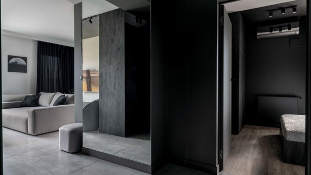 A modern, minimalist room with a grey sofa, a small ottoman, and a bed with a dark headboard. - AS HOME Niegaleria (Wrocław)