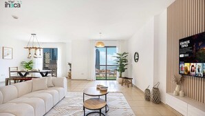 Ein modernes Wohnzimmer mit einer Couch, einem Couchtisch und einem Fernseher, der 'Stranger Things' zeigt. Es gibt einen Balkon mit Blick auf Gebäude und eine Pflanze im Raum.