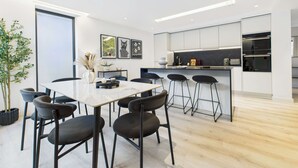 A modern kitchen with white cabinets, a central island with bar stools, and a dining area with a table and chairs. - Edgy & Modern Balcony Apartment by Borough Market (Greater London)