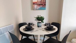 Een eetruimte met een ronde tafel gedekt voor twee personen, zwarte stoelen, een witte tafelkleed en een stadsgezichtschilderij aan de muur.